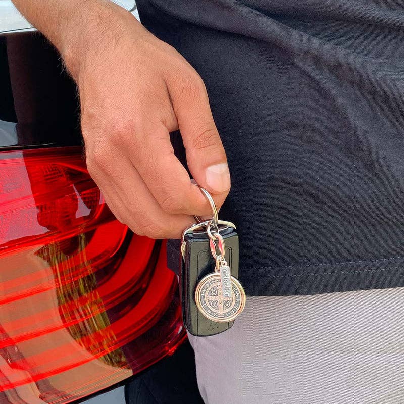 A man wearing a black shirt and gray bottoms leaning on a black car against the taillight, holding a black key fob that also has a silver St. Benedict medal on the keychain.