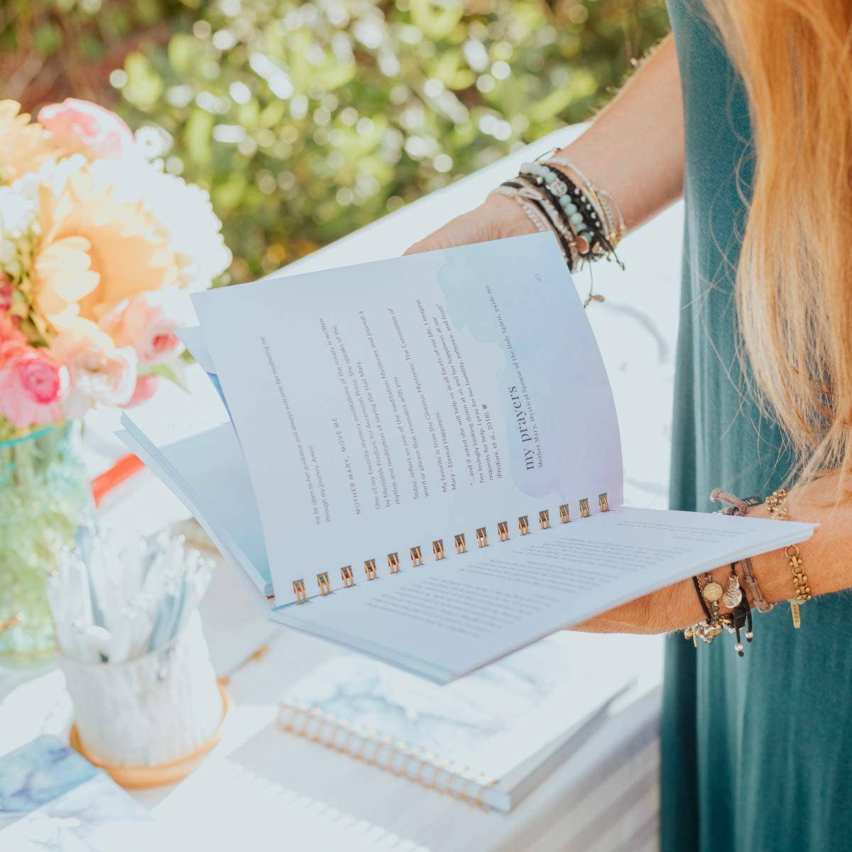 A lady holding an open page of a 40-Day Devotional to Holy Habits, "Mother Mary, Mother Me" compiled by Amy D'Ambra and Adina Braatz from My Saint My Hero. The lady is standing, wearing a teal dress and bracelets, standing over a table with a floral arrangement. She is outside.