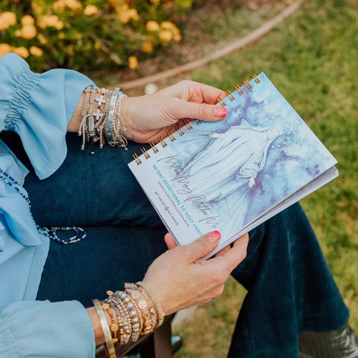 A lady holding a 40-Day Devotional to Holy Habits, "Mother Mary, Mother Me" compiled by Amy D'Ambra and Adina Braatz from My Saint My Hero. The lady is wearing gold and silver bracelets, blue jeans and a blue shirt. She is sitting on a chair outside on the grass with flowers nearby.