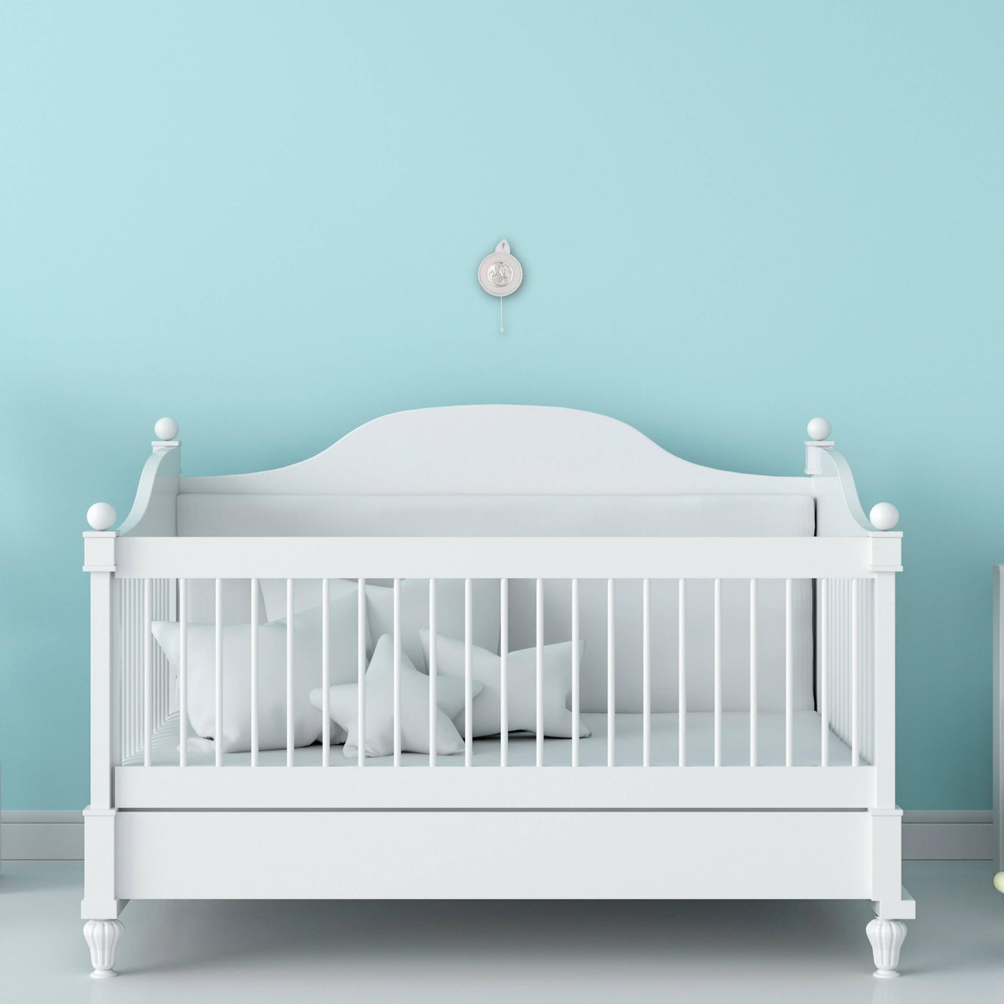 A White Round Baby Crib Medal featuring a guardian angel in silver, hanging on a white ribbon and rope, hanging on a blue wall above a white baby crib.