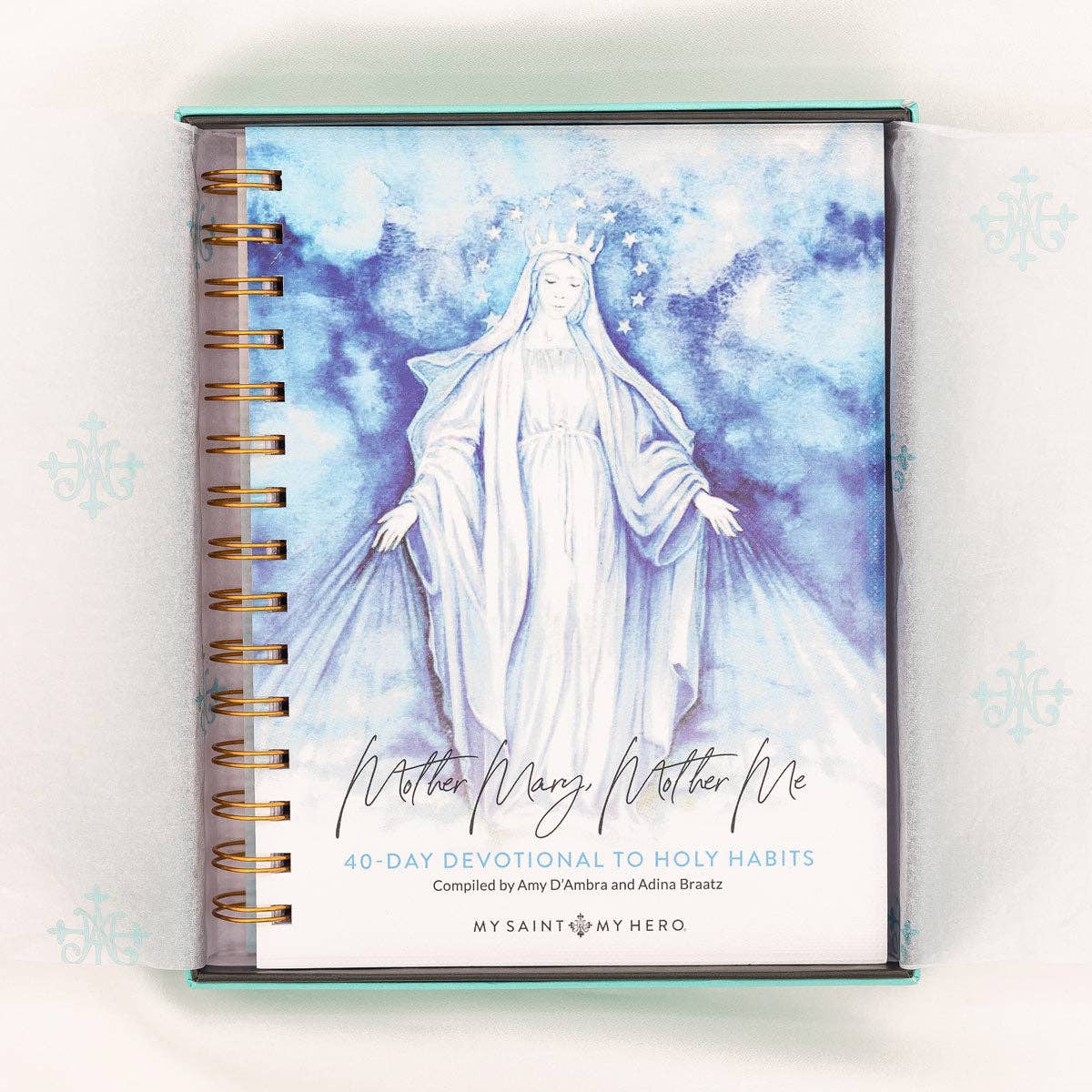 A 40-Day Devotional to Holy Habits, "Mother Mary, Mother Me" compiled by Amy D'Ambra and Adina Braatz from My Saint My Hero. The devotional is a spiral bound book with Mother Mary on the cover in a white image on a blue cloud background.