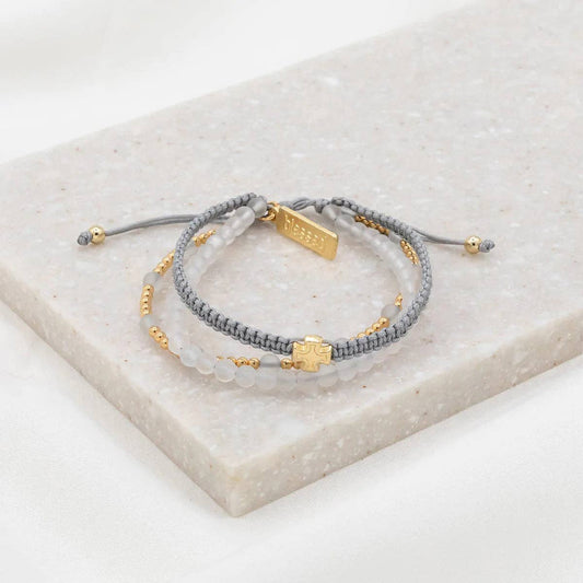 Gray Frosted White, and Gold triple pull bracelet featuring a gold cross, and gold "blessed" tag, sitting on a stone.