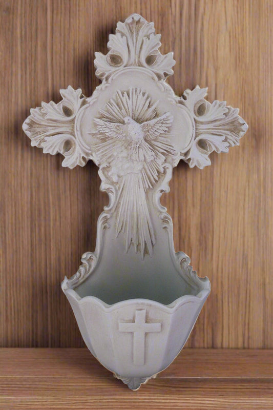 An ornate Holy Water cross font featuring a dove as the depiction of the Holy Spirit with rays around the dove.  At the bowl of the font, there is a plain embossed cross. Displayed in front of a wood background.