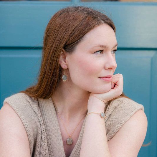 A lady wearing Saint Benedict Medal earrings in a gray design on gold hanging earrings. The lady is wearing a brown shirt, looking to the side away from the camera, in front of a blue background, sitting with her hands against her chin.
