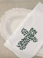 A hand towel featuring a cross out of greenery, folded, and placed on a white plate sitting on a linen tablecloth.