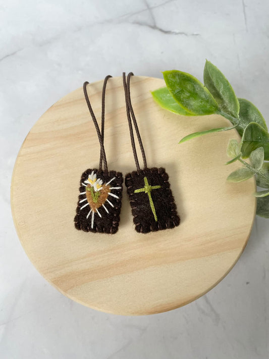 Brown Scapular featuring a green cross and brown heart with white flowers in the center and rays around the heart.